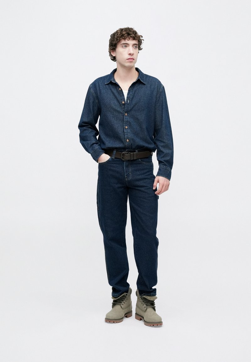 Denim shirt in dark blue with button front and collar, paired with dark blue jeans and olive green boots featuring laces and textured soles.