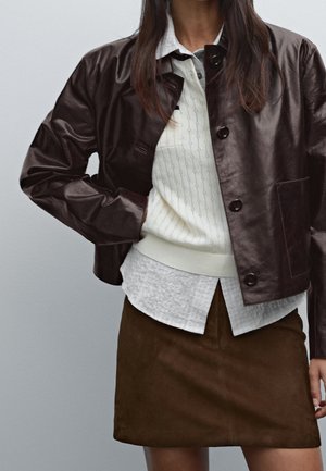 Femme portant une veste en cuir marron, un pull en maille crème par-dessus une chemise blanche à col, et une jupe en daim marron, debout devant un fond uni.