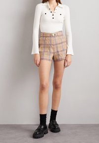 Haut blanc à manches longues côtelé avec col à boutons ; shorts ajustés marron à carreaux avec poches et ceinture ; mocassins noirs épais avec chaussettes.