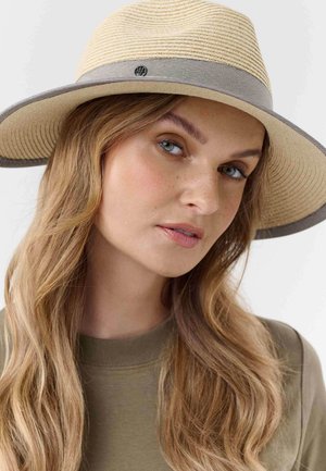Woman with long blonde hair wearing a beige wide-brimmed hat with a gray band and an olive green top, looking slightly to the side.