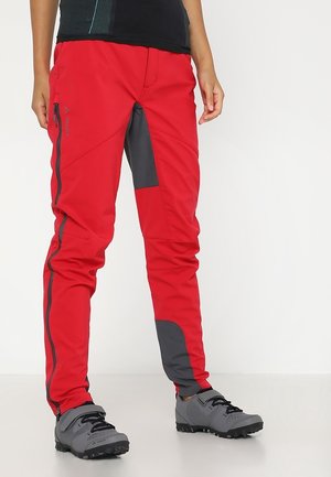 Personne portant un pantalon de plein air rouge et gris avec des zips latéraux et des chaussures de randonnée grises sur fond blanc.