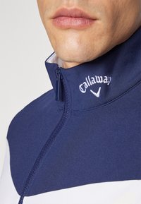 Marineblå hettejakke med hvite detaljer; har høy krage og brodert "Callaway" logo i hvitt. Glatt tekstur og tettsittende design.