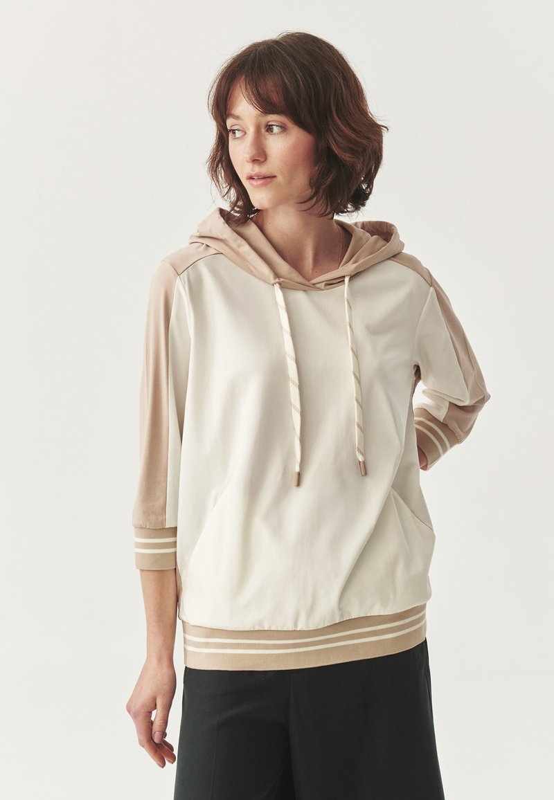 Sudadera con capucha en crema y marrón, con mangas de tres cuartos, cordones y detalles a rayas en los puños y el dobladillo. Textura de tela suave.