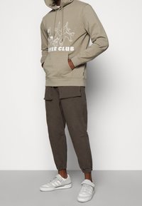 Beżowa bluza z kapturem z białym napisem "COFFEE CLUB", zestawiona z ciemnobrązowymi spodniami cargo. Białe sneakersy z tekstylnymi i gumowymi akcentami.