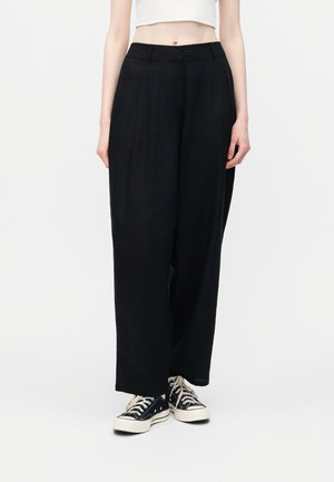 Personne portant un pantalon noir ample taille haute avec poches latérales et des baskets basses noires à lacets et semelles blanches, se tenant sur un fond blanc.