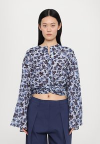Floral print blouse in light blue, featuring long bell sleeves, a knotted front, and a half-button placket. Paired with navy high-waisted pants.