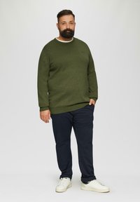 Donkergroene trui met een ronde hals, gecombineerd met marineblauwe broek en witte sneakers. De trui heeft een gestructureerde afwerking en een comfortabele pasvorm.