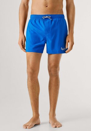 Hombre vistiendo pantalones cortos de baño azul brillante con cordón y logo de "Pepe Jeans" en la pierna izquierda, de pie descalzo contra un fondo sencillo.