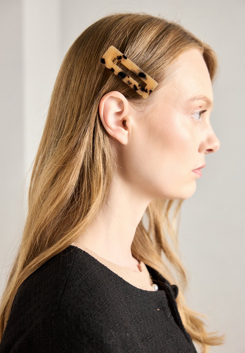A rectangular hair clip made of translucent beige acrylic with black speckles, secured in straight blonde hair. The model wears a black top.