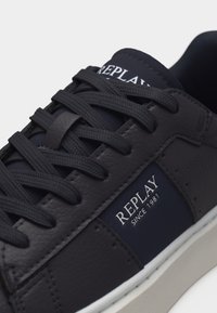 Κοντινή λήψη μαύρου sneaker Replay με υφή δέρματος, μαύρα κορδόνια και λευκό κείμενο λογότυπου που γράφει "REPLAY SINCE 1981" στην πλευρική επιφάνεια.