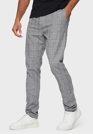 Man som bär slim-fit grå rutig byxor och vita sneakers, med ena handen i fickan, står mot en ljusgrå bakgrund.