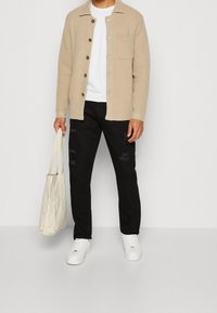 Chaqueta de camisa texturizada beige con botones marrones, camiseta blanca, pantalones negros desgastados y zapatillas blancas. Lleva una bolsa tote crema.
