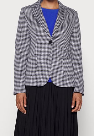 Kvinde iført en sort-hvid hundetandsmønstret blazer over en blå top og et sort plisseret skørt, stående mod en ensfarvet baggrund.