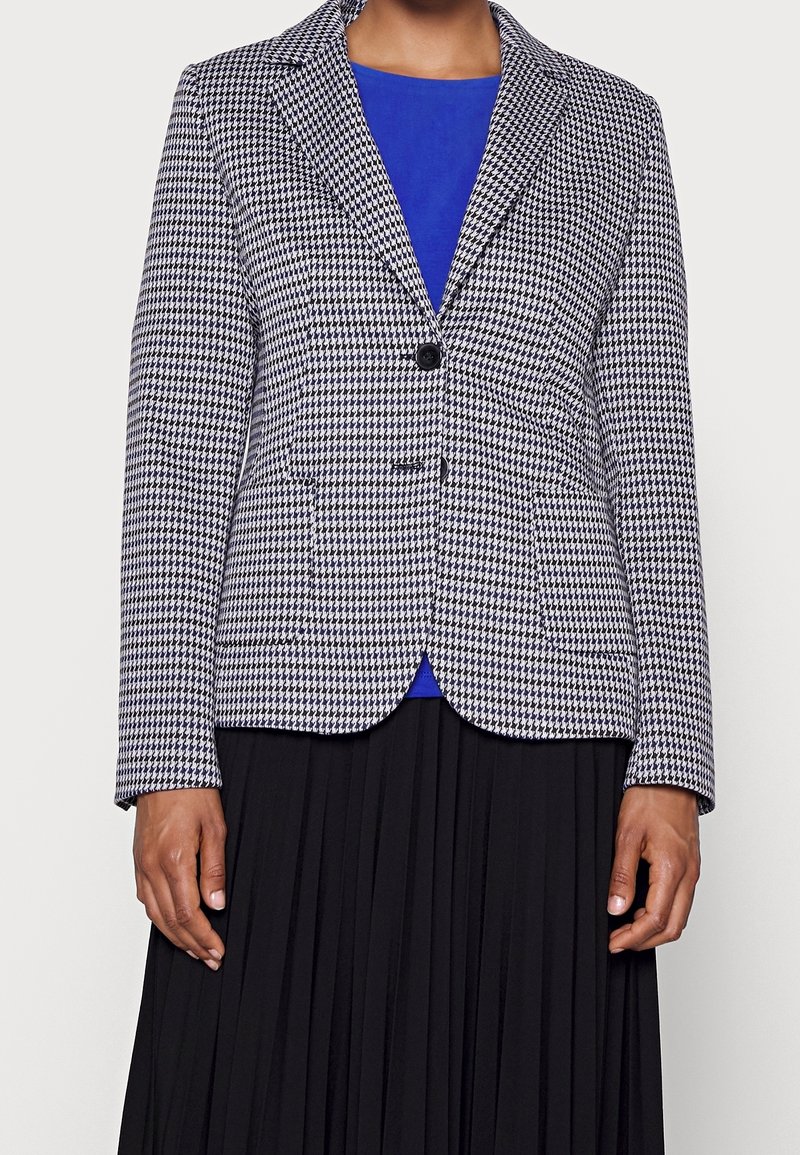 Femme portant un blazer pied-de-poule noir et blanc sur un haut bleu et une jupe noire plissée, debout devant un fond uni.
