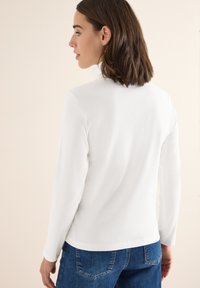 Biała bluzka z długim rękawem o prążkowanej fakturze i wysokim dekolcie, zestawiona z niebieskimi dżinsami. Widoczne detale przeszycia.
