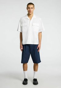 Hvid kortærmet skjorte med knapper og to frontlommer, parret med mørkeblå knælange shorts, hvide sokker og sorte sko.