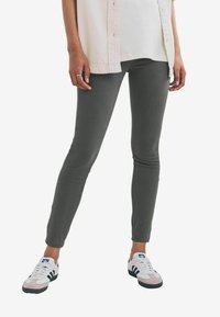 Grijze stretchy leggings met een slim fit, gecombineerd met een lichtgekleurde overhemd met knopen en witte sneakers met blauwe en zwarte accenten.