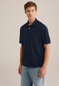 Temno modra polo majica iz bombaža, s spiščenim gumbom, kratkimi rokavi in prsno žeppo z diskretnim logotipom.