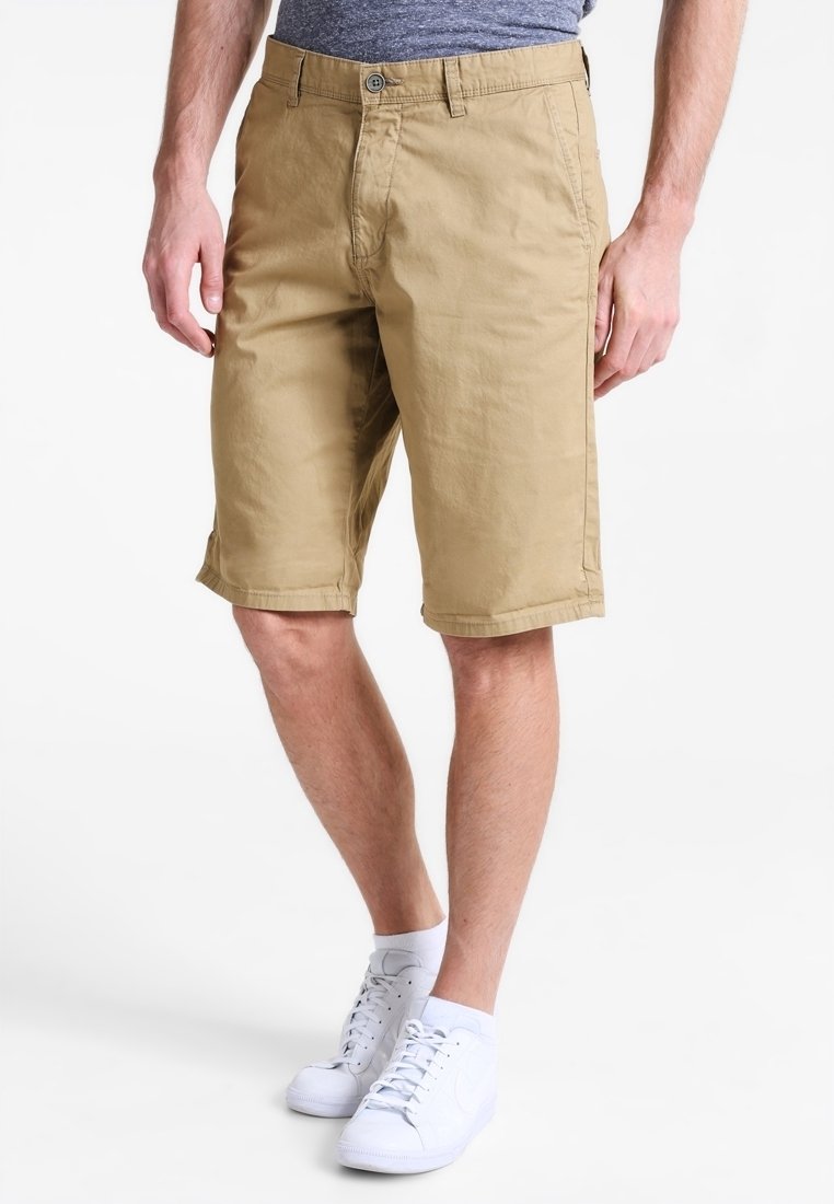 Homme portant un short beige longueur genou, une chemise grise, des chaussettes blanches basses et des baskets blanches, debout devant un fond blanc uni.
