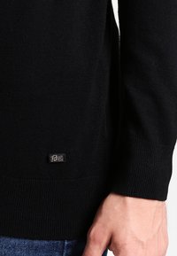 Svart stickad tröja med ribbade ärmslut och nederkant, med en liten, silverfärgad varumärkesetikett längst ner till vänster. Texturerad yta.