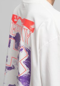 T-shirt en coton blanc avec de grands imprimés graphiques rouges et violets, comportant le texte "EDINBURGH" et des formes accentuées sur la manche.