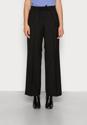 Personne portant un pantalon noir à jambes larges avec une taille élastique et des bottines noires, debout devant un fond blanc uni.