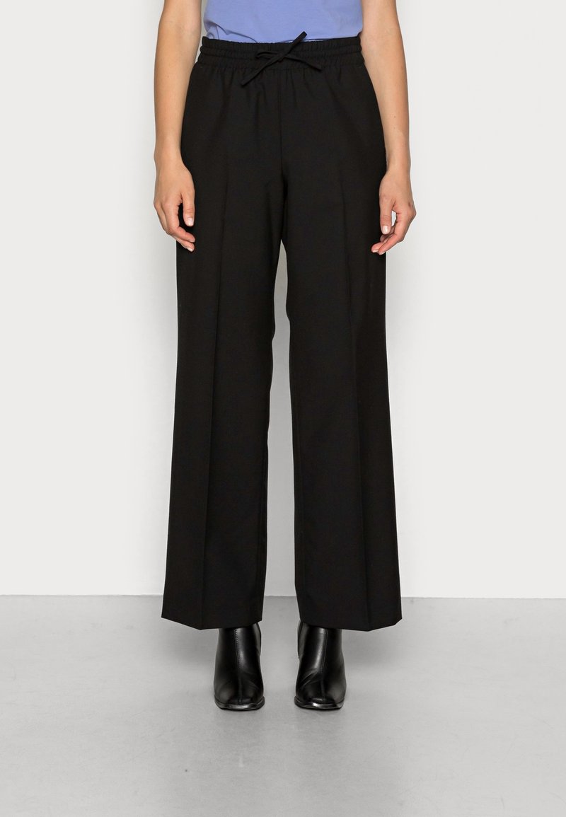 Personne portant un pantalon noir à jambes larges avec une taille élastique et des bottines noires, debout devant un fond blanc uni.