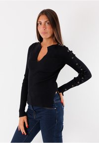 Top lungo maniche nere a coste con scollatura a V, caratterizzato da dettagli metallici ad occhielli sulle maniche e fessure laterali, abbinato a jeans blu.