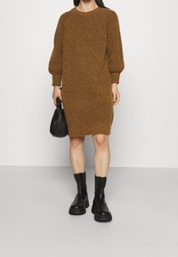 Brun stickad klänning med långa puffärmar; bärs med svarta ankelboots och en liten svart handväska. Enkel, rak skärning och strukturerad tyg.