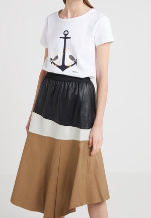 Vrouw draagt een wit shirt met korte mouwen met een print van een marineblauwe anker en meeuwen, gecombineerd met een rok in kleurvlakken in zwart, wit en bruin.