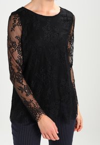 Top in pizzo nero con maniche lunghe trasparenti, motivo floreale, scollatura rotonda e vestibilità comoda, abbinato a pantaloni navy a righe sottili.