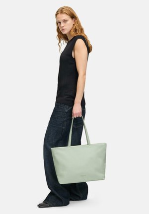 Hellgrüne Leder-Umhängetasche mit zwei oberen Tragegriffen, rechteckiger Form, glatter Textur, gehalten von einer Person, die ein schwarzes ärmelloses Oberteil und Jeans trägt.