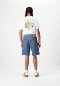 T-shirt blanche avec un design floral vert et orange au dos, associée à un short en denim bleu, des chaussettes et des chaussures marron clair.