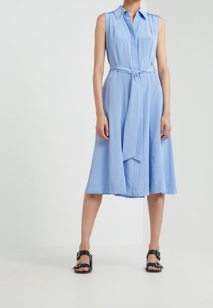 Mouwloze lichtblauwe midi-jurk met kraag en tailleband, gedragen met zwarte sandalen met hak en gespen.