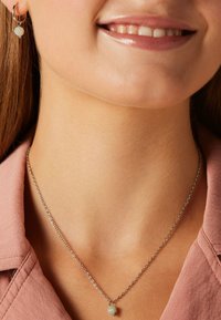 Lächelnde Person, die eine silberne Halskette mit einem kleinen sechseckigen Anhänger und passenden silbernen Kreole mit einer Perle trägt.