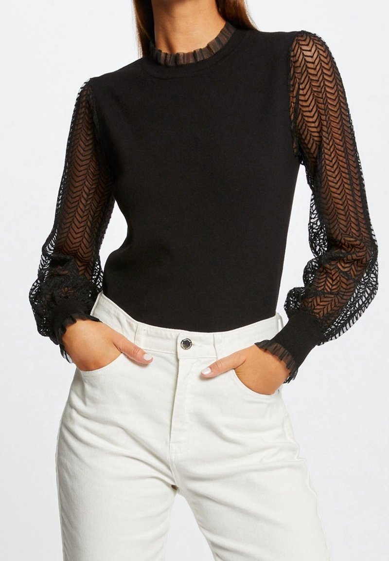 Haut noir ajusté avec manches longues en tissu transparent à motif et poignets à volants, associé à un pantalon blanc taille haute, mains dans les poches.