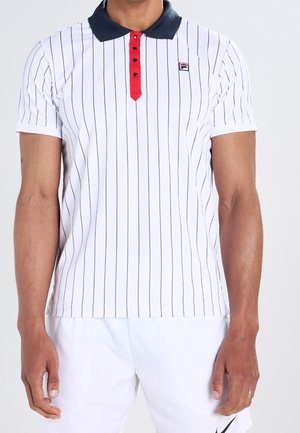 Mann trägt weißes Poloshirt mit dünnen schwarzen vertikalen Streifen, marineblauem Kragen, roter Knopfleiste und kleinem Logo auf der Brust, kombiniert mit weißen Shorts.