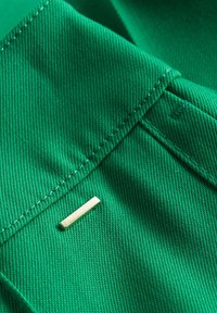 Groene stof met een gladde textuur en zichtbare stiksels, met een kleine rechthoekige metalen accent dat near de rand is ingebed.