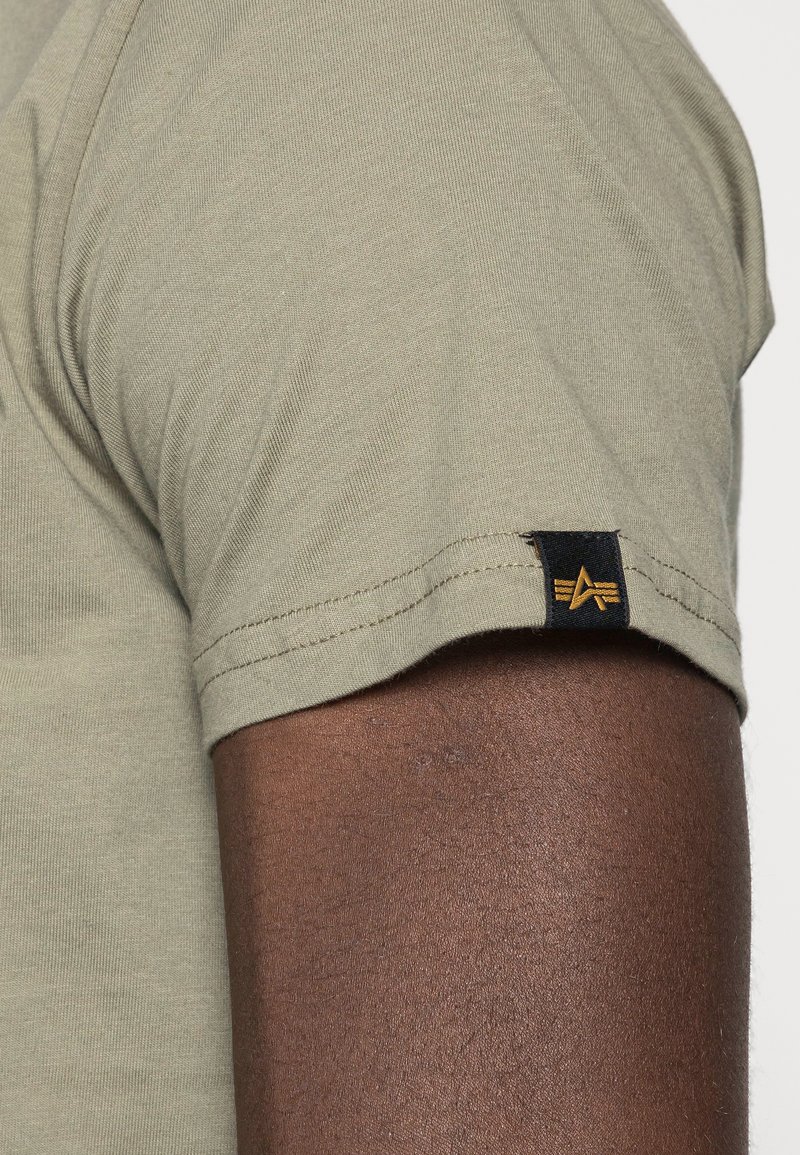 Camiseta de manga corta verde oliva con una textura suave y una etiqueta negra que presenta un logo dorado en la manga. La costura está bien confeccionada.