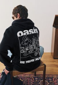Zwarte hoodie met een grote witte afbeelding op de achterkant, die een scène toont met bomen, mensen en voertuigen, met de tekst "oasis" en "BE HERE NOW."