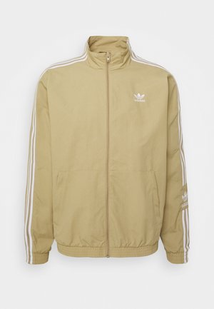 Veste de survêtement beige Adidas avec des bandes blanches sur les manches, fermeture éclair intégrale à l'avant, poignets et ourlet élastiques, et logo Adidas sur la poitrine et la manche.