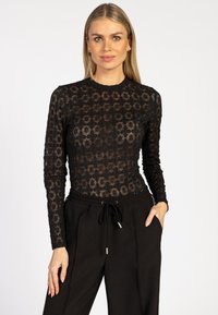 Vrouw met lang blond haar draagt een zwart met bloemenkant langetermijn top en zwarte broek met trekkoord, staande tegen een witte achtergrond.