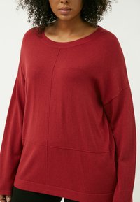 Maglione rosso oversize con scollatura rotonda, maniche lunghe e una consistenza morbida. Presenta un dettaglio di cucitura sulla parte anteriore e laterale.