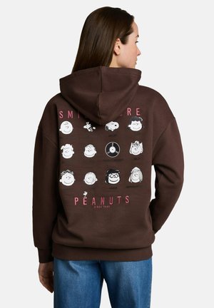 Kobieta w brązowej bluzie z kapturem z nadrukowanymi na plecach twarzami i imionami postaci z Snoopy i Peanuts, w połączeniu z niebieskimi dżinsami.