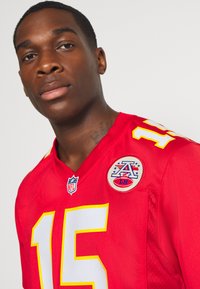 Piros NFL mez fehér "15" számmal az elülső részen, sárga díszítésekkel és hímzett folttal, amely csillag- és focilabdadesign-t tartalmaz a vállán.