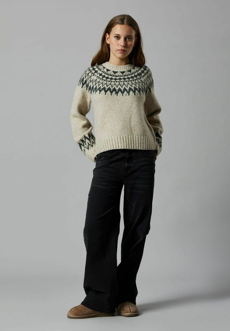 Suéter de punto en beige con un yugo gris oscuro estampado. Presenta puños y dobladillo acanalados. Combinado con jeans oscuros y calzado marrón.