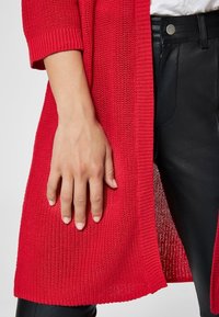 Lång röd stickad cardigan med texturerad yta, korta ärmar och öppen framsida, bärs över svarta läderbyxor.
