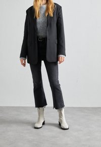 Blazer negro sobre una camiseta de punto gris, acompañado de jeans oscuros de campana y botas de tobillo de ante blancas. Detalles destacados incluyen el dobladillo sin acabado en los jeans.