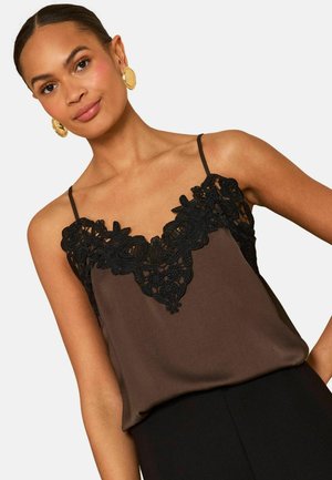 Frau trägt ein braunes ärmelloses Oberteil mit schwarzer, blumiger Spitzenverzierung am Ausschnitt und goldenen runden Ohrringen, posiert mit einem leichten Lächeln.