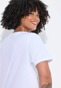Bílé bavlněné tričko s krátkými rukávy a kulatým výstřihem. Hladká textura a volný střih. Na ruce je viditelné tetování poblíž lokte.